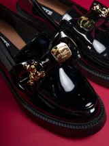 Mocasín de charol negro con plataforma, detalle de lazo y aplique de oso dorado.
