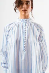 Blusa de algodón blanco con diseño de rayas verticales y textura de pequeños puntos en relieve. Presenta cuello alto con volados, cierre frontal con botones forrados y mangas largas abullonadas con puños ajustados.