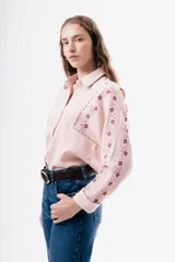 Camisa de algodón color rosado con cuello clásico y cierre frontal de botones. Presenta detalles de bordados florales en tonos rojizos a lo largo de las mangas y costuras con encaje decorativo en los hombros.
