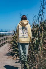 Hoodie amarillo con capucha, de corte holgado y diseño de espalda con bordado gráfico de olas, sol y la inscripción 'Glassy Waves'. Presenta detalles de ondas bordadas en el ruedo inferior.