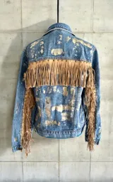 Campera de jean azul intervenida con detalles en tono dorado pálido, con flecos de cuero beige en la espalda y las mangas, decorados con tachas metálicas.