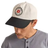 Gorra de béisbol color beige con visera negra y parche circular con diseño de flor roja y texto en negro.