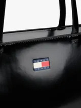 Bolso tipo tote de Tommy Jeans en color negro con acabado brillante. Presenta un diseño minimalista con doble asa superior y el logo distintivo de la marca aplicado en la parte frontal.
