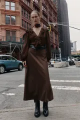 Vestido midi de color marrón con diseño combinado, escote en pico con volantes, mangas largas con puños elásticos y falda evasé. Incluye un cinturón negro con hebilla dorada en la cintura y cierre lateral con cremallera oculta.