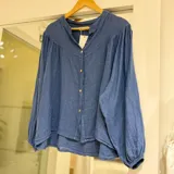 Camisa de lino color azul jean, con cuello mao, cierre frontal con botones y mangas largas abullonadas con puños elásticos.