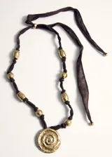 Collar con cinta marrón ajustable en la parte posterior, abalorios dorados texturizados a lo largo de la tira y un dije circular central dorado con diseño de espiral en relieve y pequeños cristales incrustados.