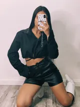 Camisa cropped negra con cuello clásico, mangas largas con puños abotonados y cierre frontal con botones. Presenta detalle de tachas plateadas en el cuello.