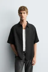Camisa negra de manga corta con cuello solapa y textura arrugada o plisada. Corte relajado (relaxed fit) con aberturas laterales y cierre frontal de botones.