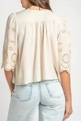 Blusa color arena de algodón peinado, con escote redondo, mangas cortas abullonadas con bordados suizos y detalles de alforcas en el frente.