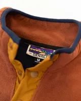 Buzo polar de hombre con diseño color block en tonos azul marino, amarillo y naranja. Cuenta con cuello alto con cierre de botones a presión, bolsillo frontal con solapa y cierre, y mangas largas con puños elásticos.