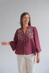 Blusa color bordó con bordados florales en tono crema en la parte superior frontal. Presenta cuello redondo con pequeña abertura, cierre frontal con botones y mangas largas acampanadas con puños ajustados.