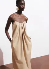 Vestido largo color beige, de diseño recto sin tirantes (palabra de honor) con escote corazón y detalle de pliegues en la parte delantera. Cuenta con cierre de cremallera en la espalda y bolsillos laterales.