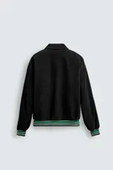 Cazadora estilo varsity confeccionada en pana de algodón azul, con interior ligeramente acolchado. Presenta cuello solapa, cierre frontal con botones a presión, bolsillos laterales de vivo y acabados en rib a rayas en puños y cintura. Incluye múltiples parches bordados decorativos en el frente.