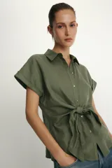 Camisa blanca confeccionada en tejido con lino, con cuello solapa y manga muy corta con terminación en vuelta. Presenta un detalle de nudo en el delantero y cierre frontal con botones metálicos.