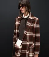 Trench coat largo de corte clásico con estampado de cuadros en tonos marrones y beige. Presenta cuello con solapas, cierre frontal con botones, mangas largas y bolsillos laterales aplicados.