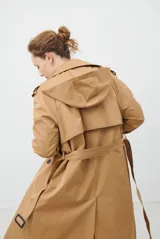 Trench coat color beige con cuello de solapa, cierre frontal abotonado, bolsillos laterales y mangas largas con cintas ajustables. Incluye capucha desmontable y forro interior rayado. La tela exterior cuenta con protección repelente a la lluvia y manchas.