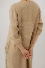 Vestido largo de lino color tostado, de manga larga, con escote redondo y diseño amplio y fresco. Cuenta con un delicado bordado en la espalda con motivos marinos y la frase "no se lo digas".