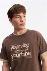 Remera marrón de manga corta confeccionada en algodón, con cuello redondo y ribetes en contraste de color tostado en el cuello y los puños. Presenta estampa frontal con la frase "your vibe attracts your tribe" y el logo de Mistral.