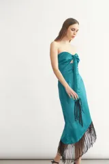 Vestido midi sin mangas color turquesa, con escote strapless y detalle de nudo frontal. El ruedo presenta un corte asimétrico con flecos negros largos.
