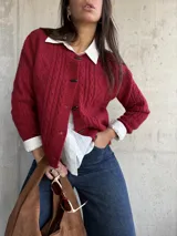 Cardigan de punto con textura trenzada en color bordó, con cuello redondo y cierre frontal mediante botones tipo alamar. Presenta mangas largas con puños acanalados y un corte clásico.