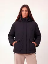 Campera negra de corte recto con capucha, cierre frontal oculto por solapa y bolsillos delanteros con solapa. Presenta un diseño minimalista con un pequeño parche circular en el pecho.