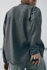 Camisa de corte oversize confeccionada en algodón, con estampado de cuadros pequeños en tonos grises. Presenta cuello solapa, cierre frontal con botones ocultos y mangas largas con puños rectos.
