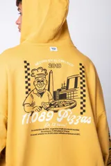 Buzo canguro amarillo ocre oversize de algodón rústico con capucha, cordones negros con letras blancas, bolsillo frontal y puños y cintura acanalados. Presenta estampados en el frente y las mangas.