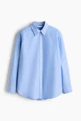 Camisa de algodón color celeste, con cuello clásico, cierre frontal de botones y mangas largas con puños sencillos. Presenta corte relajado y bajo redondeado.