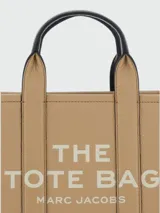 Cartera tipo tote bag Marc Jacobs en color tostado, con detalles en negro y blanco. Cuenta con las inscripciones "THE TOTE BAG" y "MARC JACOBS" estampadas en el frente. Posee doble asa de mano superior y correa de hombro ajustable y desmontable.