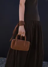 Bolso de mano tipo bowling de cuero gamuzado color marrón, con dos asas cortas y detalle de tiras con nudo y flecos colgantes en un lateral. Cierre superior con cremallera.
