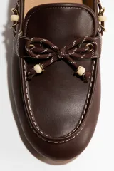 Mocasines estilo náutico de color marrón, con cordones decorativos bicolores y cuentas claras en la parte frontal y laterales. Cuenta con costuras pespunteadas en contraste y suela plana.