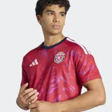 Camiseta de fútbol Adidas de la selección de Costa Rica, modelo Primera Equipación 26. Diseño principal en rojo con estampado abstracto en tonos morados/rosados. Presenta cuello redondo en azul marino y las tres bandas de Adidas en los hombros en blanco. Incluye el escudo de la federación costarricense.