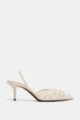 Zapato de tacón destalonado en color crudo, con cuerpo de mesh transparente con bordados florales y aplicación de abalorios brillantes. Cuenta con tira trasera elástica, tacón medio fino y acabado en punta.