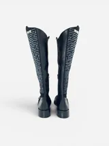 Bota de caña alta en cuero negro con punta redonda y taco bajo. Presenta un panel lateral de gamuza con bordado geométrico en hilo gris, herraje metálico decorativo en el empeine y cierre interno. El diseño cuenta con una boca asimétrica y forro interior de cuero.