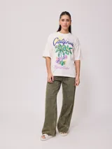 Remera color amarillo pastel con estampado de palmeras, flores de hibisco y texto 'California Ride the Tide Rusty Tribe'.