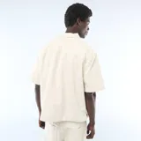 Camisa vaquera de manga corta en color beige, con cuello clásico, cierre frontal de botones y un bolsillo aplicado en el pecho.