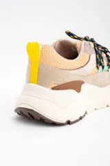 Zapatilla urbana de diseño chunky, con paneles combinados en tonos beige, arena y gris. Presenta cordones jaspeados en azul y amarillo, tirador trasero amarillo y suela de plataforma dentada en color crema.