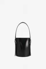 Bolso de mano tipo cubo color negro, de diseño estructurado y cilíndrico, confeccionado en cuero vacuno con acabado brillante. Cuenta con asa superior fija y correas laterales decorativas con hebillas metálicas plateadas.