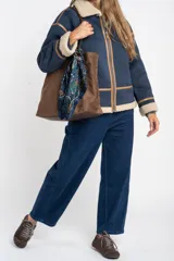 Campera tipo gamulán de corte oversize, color azul marino con interior y cuello de corderito en tono beige. Presenta detalles de costuras y bordes en cuero sintético color marrón, cierre frontal y mangas largas.