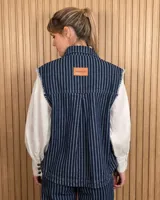 Chaleco de denim color marrón, confeccionado en 100% algodón, con bolsillos frontales amplios, cierre de botones y costuras decorativas bordadas en blanco en los bordes y los bolsillos.