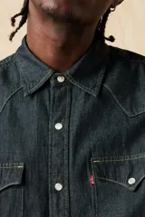 Camisa de denim azul oscuro con corte clásico, mangas largas, cierre con botones a presión, dos bolsillos frontales con solapa y costuras decorativas en los hombros.