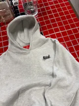 Buzo con capucha de color gris melange, con diseño estampado en la espalda que incluye el logo de la marca y elementos gráficos como un reloj y una botella. Presenta un corte holgado y puños elásticos.