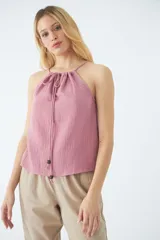 Blusa sin mangas color rosado, confeccionada en tela con textura arrugada. Presenta un escote halter con cordón ajustable que se anuda en el cuello y cae hacia el frente, decorado con cuentas de madera en los extremos.