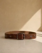 Cinto de cuero marrón claro, con hebilla metálica rectangular plateada y detalles de tachas y apliques geométricos en los extremos.