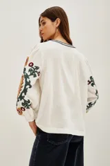 Camisa blanca de doble gasa de algodón, corte amplio y mangas largas abullonadas. Presenta bordados a mano en mangas y escote con motivos florales en verde, amarillo y violeta.