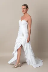 Vestido de novia blanco, sin mangas, con escote corazón y espalda en V. Confeccionado en tafeta drapeada con bordados blancos. Falda con volados y tajo frontal.