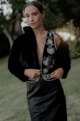 Chaleco negro con escote en V profundo, confeccionado en tela con bordados florales en hilo plateado. Presenta un diseño estructurado con ribetes plateados en los bordes y cierre frontal.