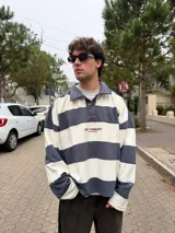 Buzo tipo polo de corte oversize con diseño de rayas horizontales anchas en color gris oscuro y crema. Presenta cuello camisero, cierre frontal parcial con botones ocultos y detalles de costuras expuestas y deshilachadas. Incluye el logo de la marca bordado en el centro del pecho.