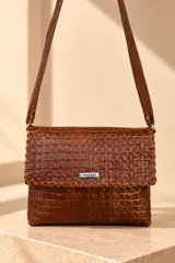 Bandolera de cuero marrón con textura símil cocodrilo, solapa frontal con detalles de costuras entrelazadas y placa metálica con el logo de la marca. Cuenta con correa larga ajustable para llevar al hombro o cruzada.