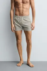 Bañador corto de hombre con cintura elástica ajustable mediante cordón blanco. Presenta un estampado geométrico repetitivo en tonos oscuros (negro/marrón oscuro) y blanco/beige. Incluye bolsillos laterales y un bolsillo trasero de vivo.